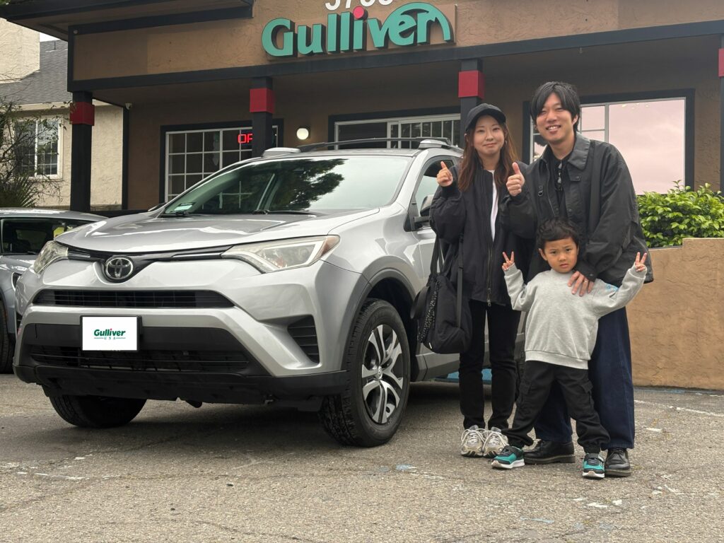 Family of three posing with a silver SUV outside a Gulliver dealership, all smiling and giving thumbs up.