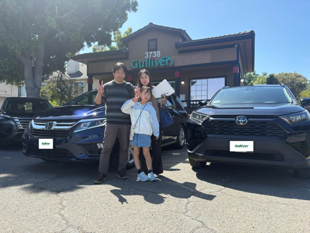 Family of three posing and making peace signs in front of a Gulliver dealership with two black SUVs under a blue sky.