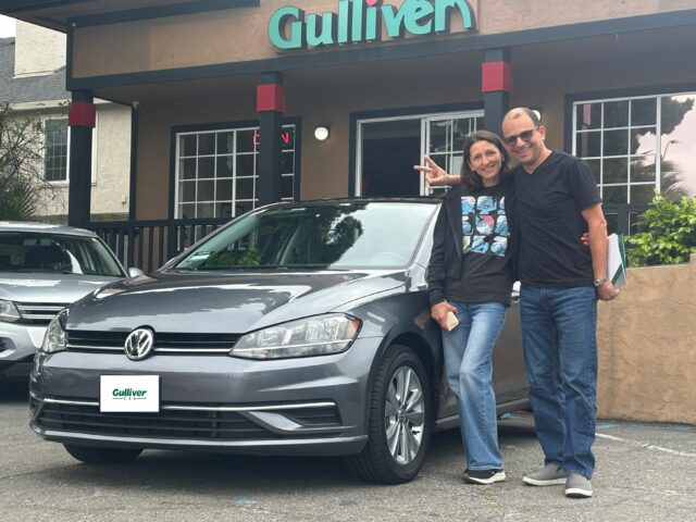 Two people stand beside a gray sedan in a Gulliver dealership parking lot, smiling at the camera.