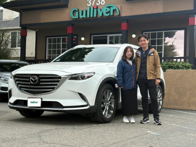 Two people smile and pose beside a white SUV in front of a Gulliver dealership storefront with teal sign and red accents in the columns.