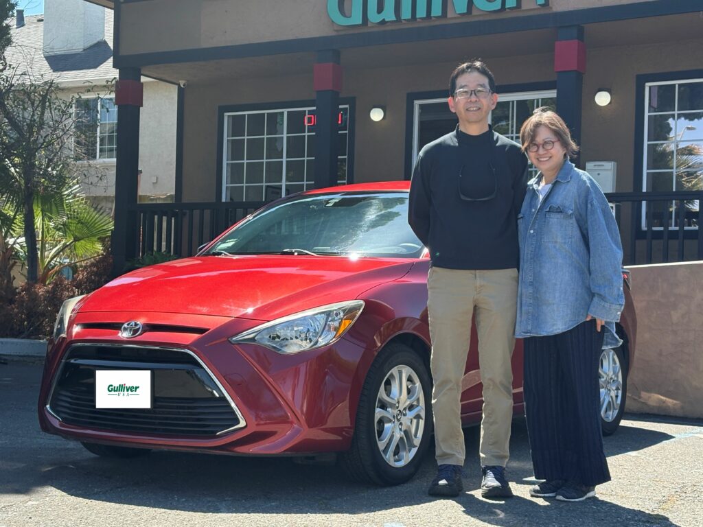 A man and a woman stand beside a red sedan in front of a Gulliver dealership, smiling at the camera.